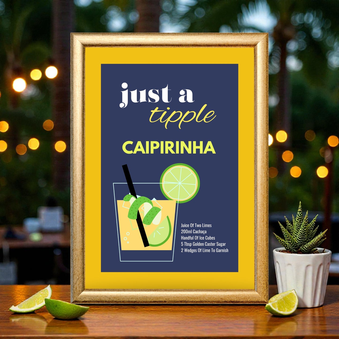 Framed caipirinha cocktail recipe print with navy and yellow design, illustrated glass with lime wedges, displayed on a wooden bar with limes, plants, and warm string lights.