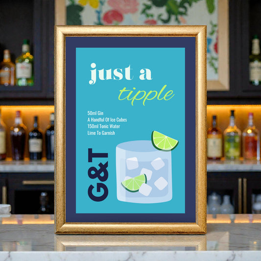 Framed ‘Just a Tipple’ gin and tonic cocktail illustration print with a blue background, shown in a gold frame on a bar counter with bottles softly blurred in the background.